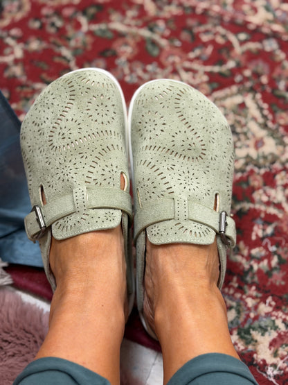 A pair of sage green slip-on shoes with laser-cut circular patterns and adjustable straps, shown on a person's feet over a red patterned rug.