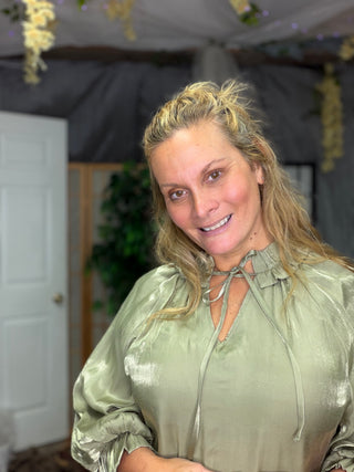 A smiling blonde woman wearing a shiny sage green satin blouse with a tie-neck detail, photographed indoors under a decorated tent or canopy.