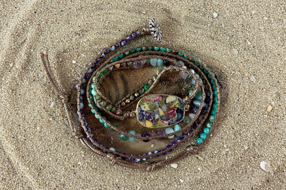 A multi-strand wrap bracelet with turquoise, purple, and brown beads, featuring a central oval floral resin charm and a silver butterfly clasp, displayed on a rustic wood slice background.
