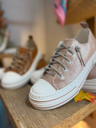 A close-up of a glittery dusty rose pink low-top sneaker with white rubber toe cap and sole, displayed on a wooden shelf.