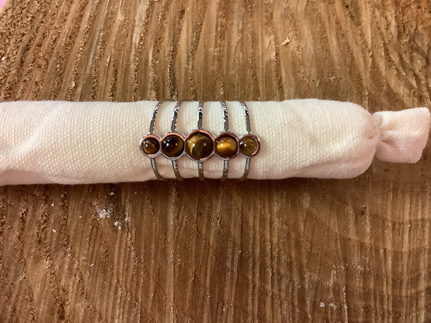 Five silver rings with round brown and amber tiger's eye stones, displayed on a white fabric sleeve over a wooden surface.