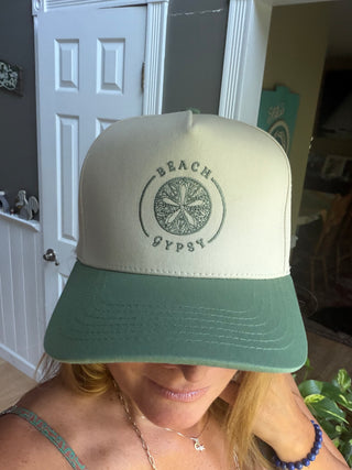 A person wearing a Beach Gypsy trucker hat with a cream-colored front panel and a sage green brim, featuring a circular sand dollar graphic on the front.