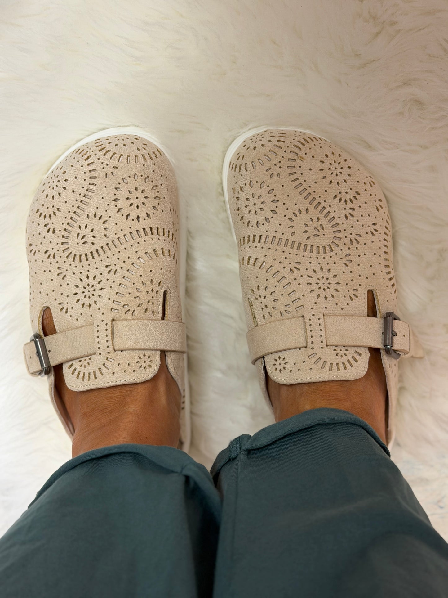 A top-down view of a person wearing beige cutout Mary Jane-style slip-on shoes with adjustable straps, paired with teal pants, standing on a white faux fur rug.