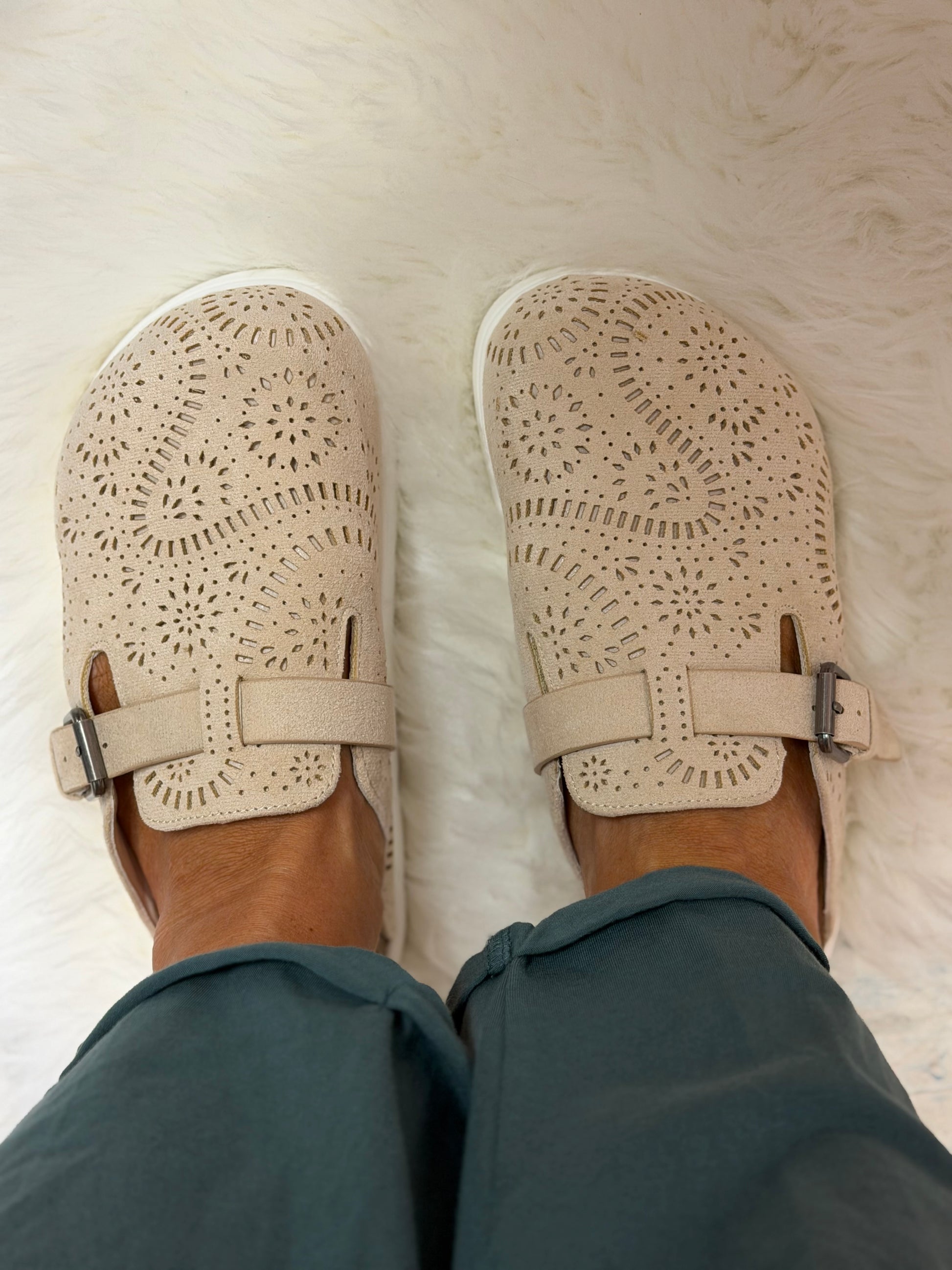 A top-down view of a person wearing beige cutout Mary Jane-style slip-on shoes with adjustable straps, paired with teal pants, standing on a white faux fur rug.