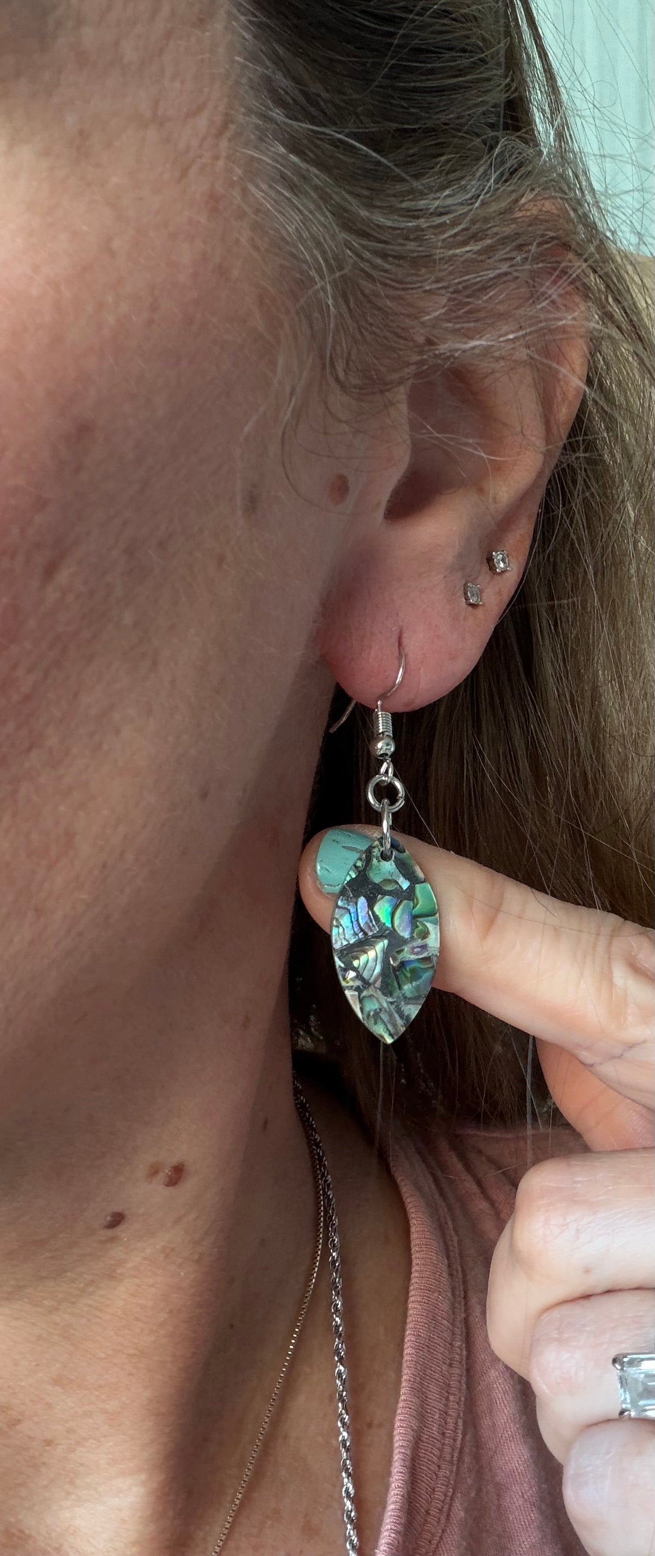 Close-up of an ear with an abalone surfboard shaped earring, with hand pointing to it.