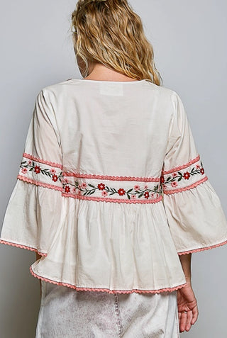 A woman viewed from the back wearing a cream-colored blouse with a floral embroidered band around the waist and flared sleeves, paired with light gray denim bottoms.