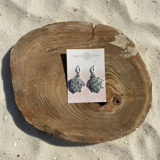 A pair of dangle earrings with green and grey stone  in a turtle shell setting, displayed on a pink Beach Gypsy card, set against a rustic wood slice on a sand background.