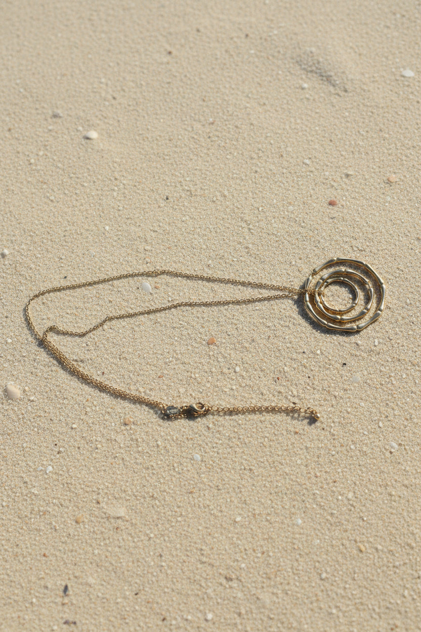A gold-toned necklace featuring a spiral pendant made of linked circular segments, resting on a sand background.