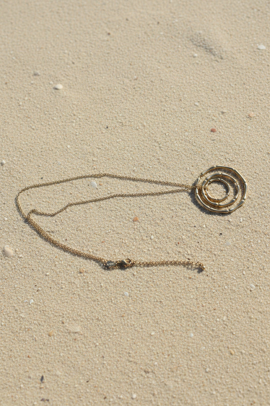 A gold-toned necklace featuring a spiral pendant made of linked circular segments, resting on a sand background.