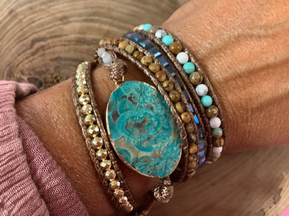 A close-up of a person's wrist wearing a bohemian-style wrapped bracelets, featuring a large oval turquoise stone centerpiece with brown and gold wraparound cords, beads, and metallic accents.