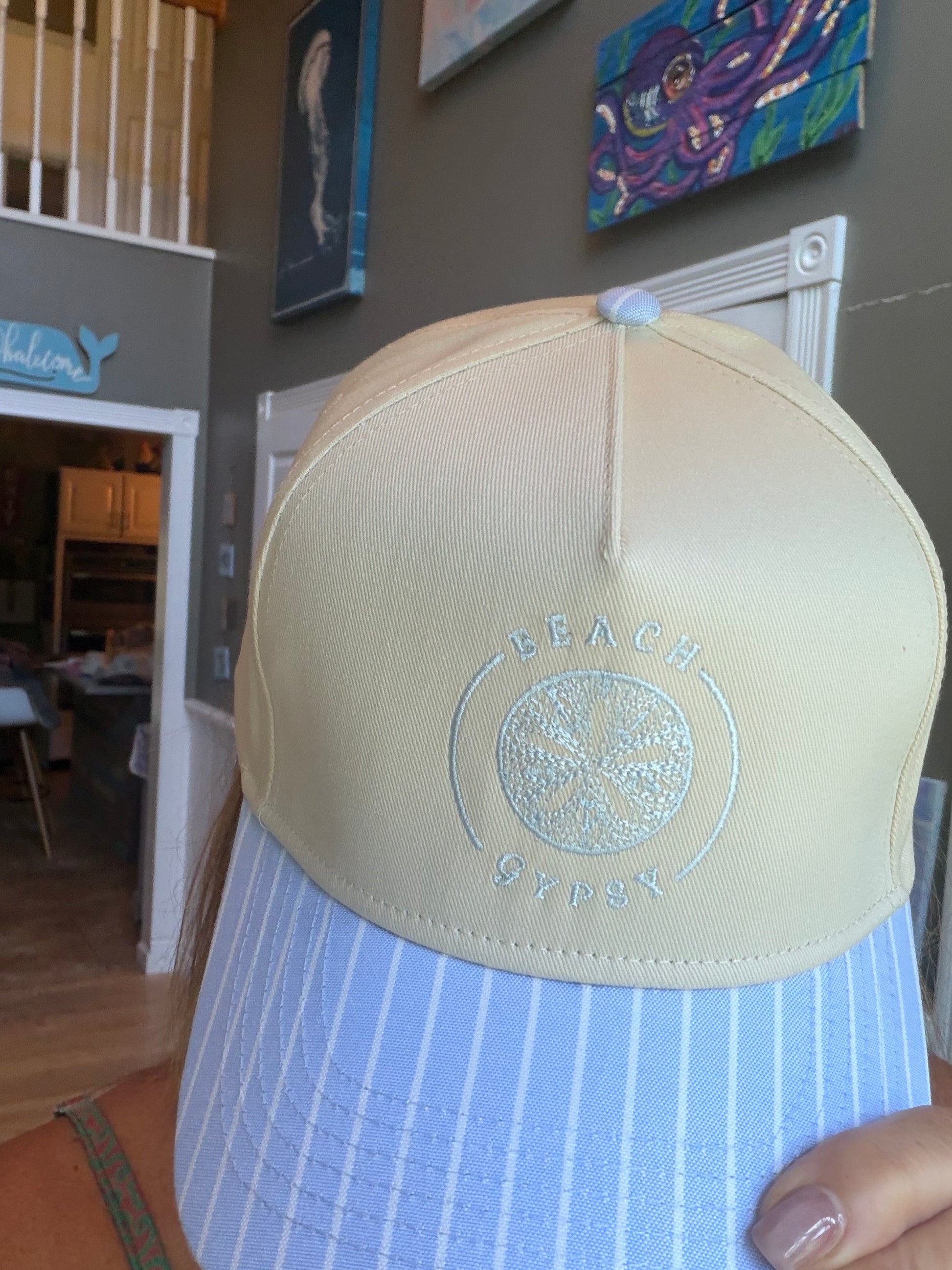 A close-up of a Beach Gypsy trucker hat with a cream-colored crown and a light blue and white striped visor, held by a person in a home setting.