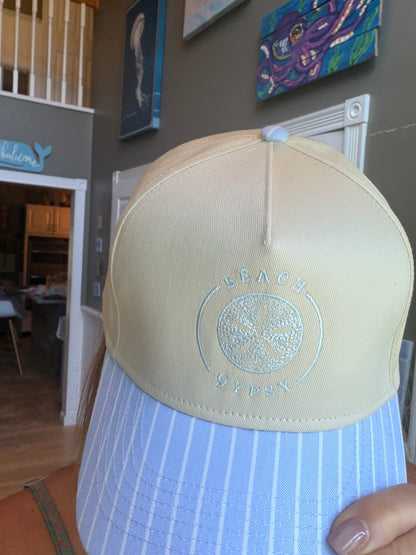 A close-up of a Beach Gypsy trucker hat with a cream-colored crown and a light blue and white striped visor, held by a person in a home setting.