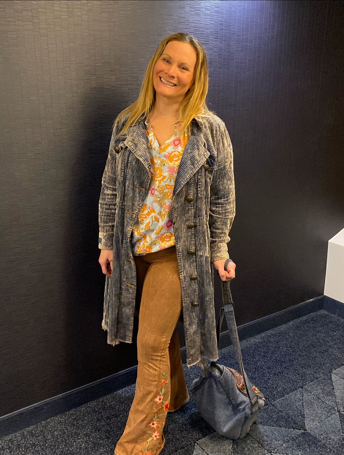 Woman standing against a black wall with a blue carpeted floor, holding a gray bag.