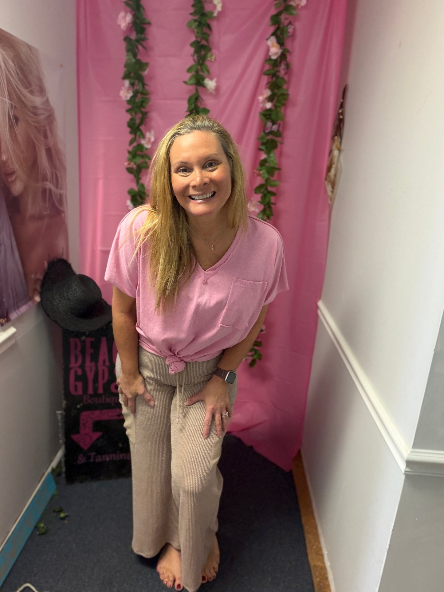 Woman in a pink shirt standing in a room with a pink curtain and posters on the wall.