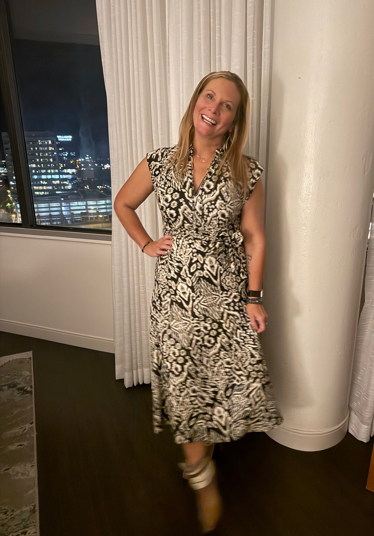 Woman in a patterned dress standing in a room with a window view of a cityscape.