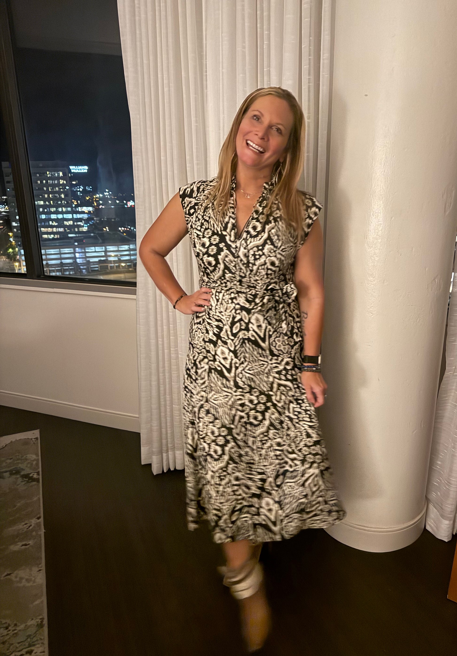 Woman in a patterned dress standing in a room with a window view of a cityscape.