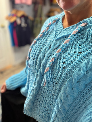 A closeup of a woman wearing a bright blue crochet knit cardigan hoodie sweater with braided hoodie Ties and dark gray pants.