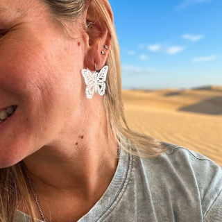 Smiling woman wearing Silver Butterfly Earrings with a gray tee, sand dunes and blue sky behind her