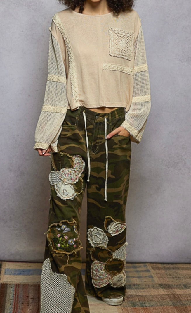 Person wearing a beige top and camouflage pants with floral patterns against a gray wall.
