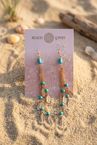 Turquoise Beaded Feather Chain Earrings with long gold-tone chains, turquoise beads and feather charms on a Beach Gypsy earring card, set in the sand with seashells and driftwood