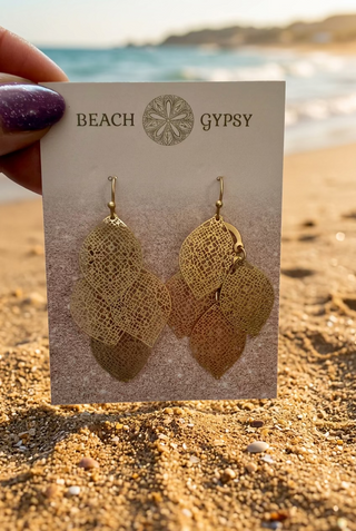 Gold-Tone Layered Filigree Leaf Dangle Earrings with overlapping lace-like leaves on a Beach Gypsy earring card, held up on the beach by the ocean