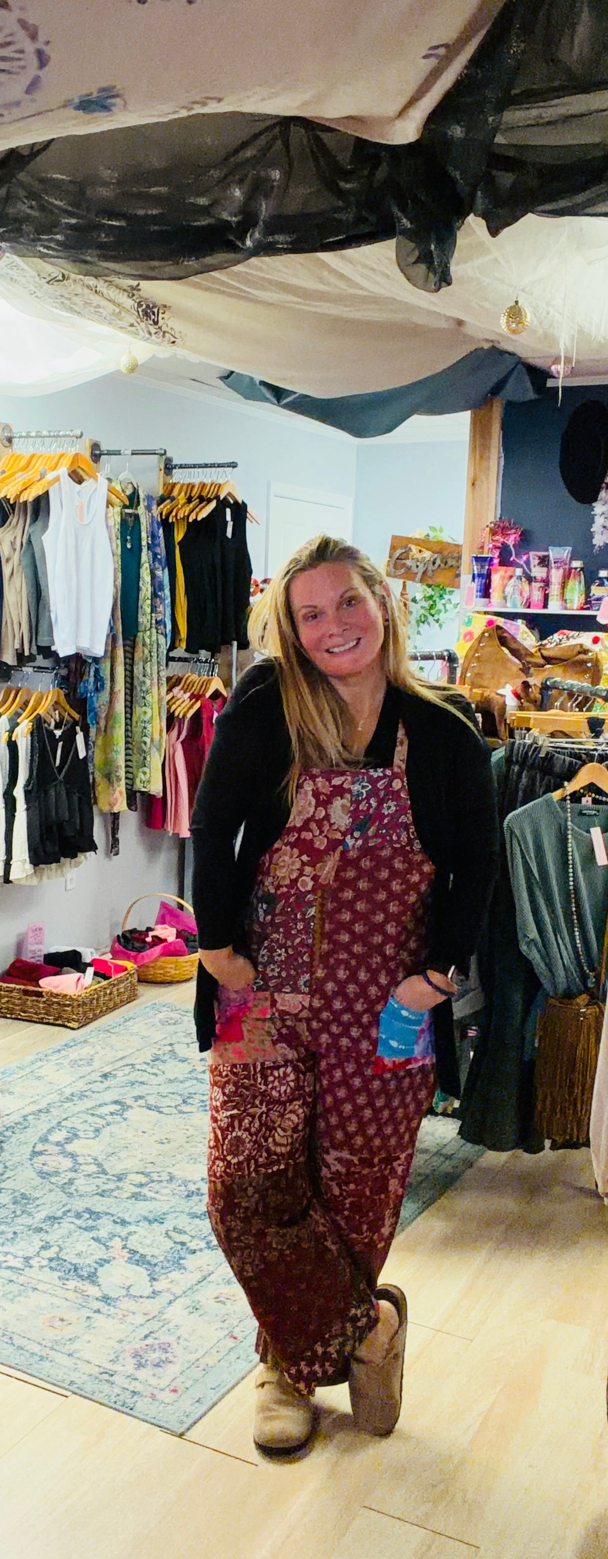Woman wearing burgundy floral overalls with black long sleeve top standing in bohemian boutique in Berlin Maryland. Beach Gypsy boutique owner showcasing boho fashion styling for women who love free-spirited clothing and unique accessories.