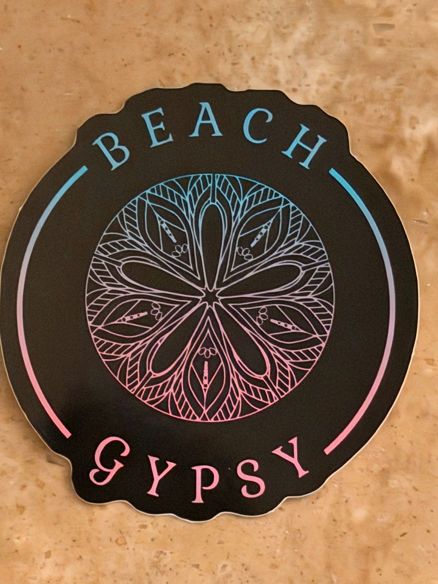 A scalloped circular sticker with a black background featuring the words 'BEACH' at the top and 'GYPSY' at the bottom, surrounding a central mandala-style floral design. The sticker is photographed on a textured tan surface.