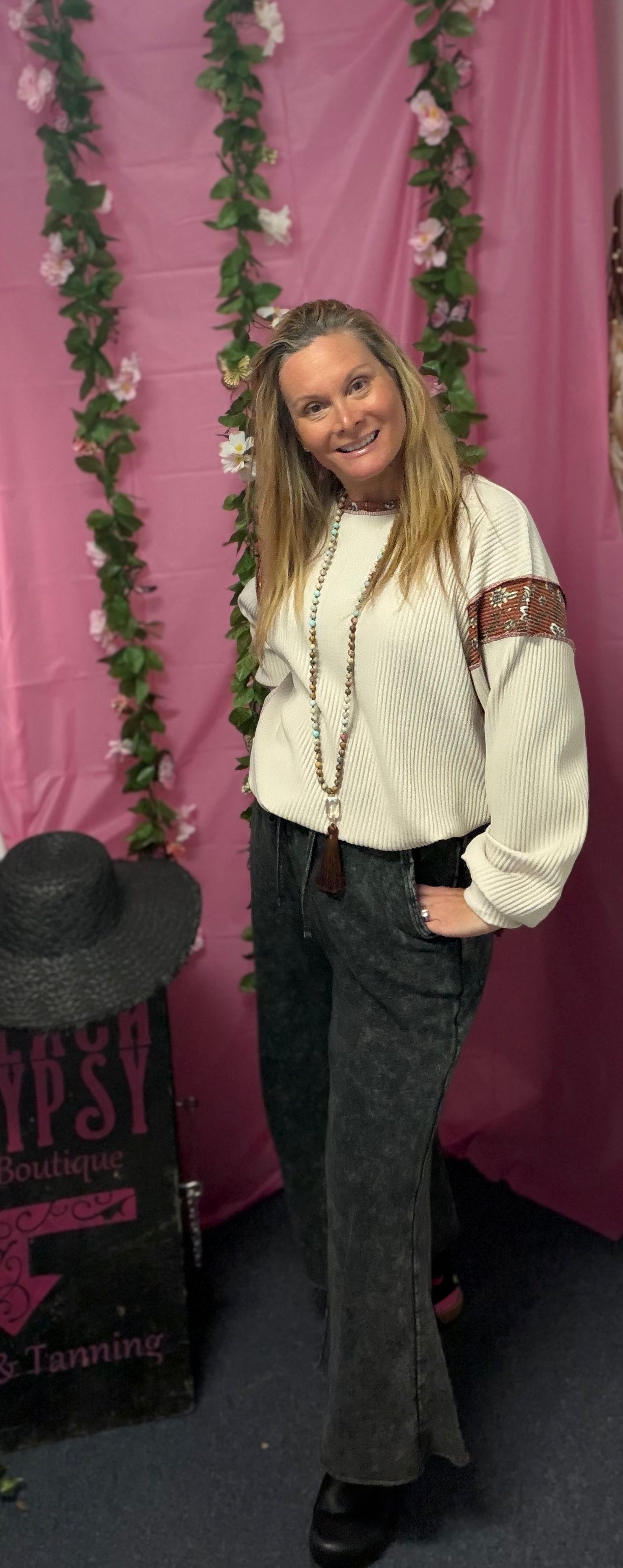 Woman posing in front of a pink backdrop with floral decorations, wearing a white sweater and dark pants.