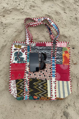 A patchwork quilted tote bag with multicolor patterns, shown on a light wood floor with a smartphone and sunglasses resting in its front pocket.