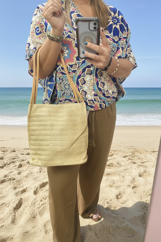A person wearing a multicolored geometric patterned short sleeve top with a V-neckline, paired with beige pants, holding a staw purse on a beach