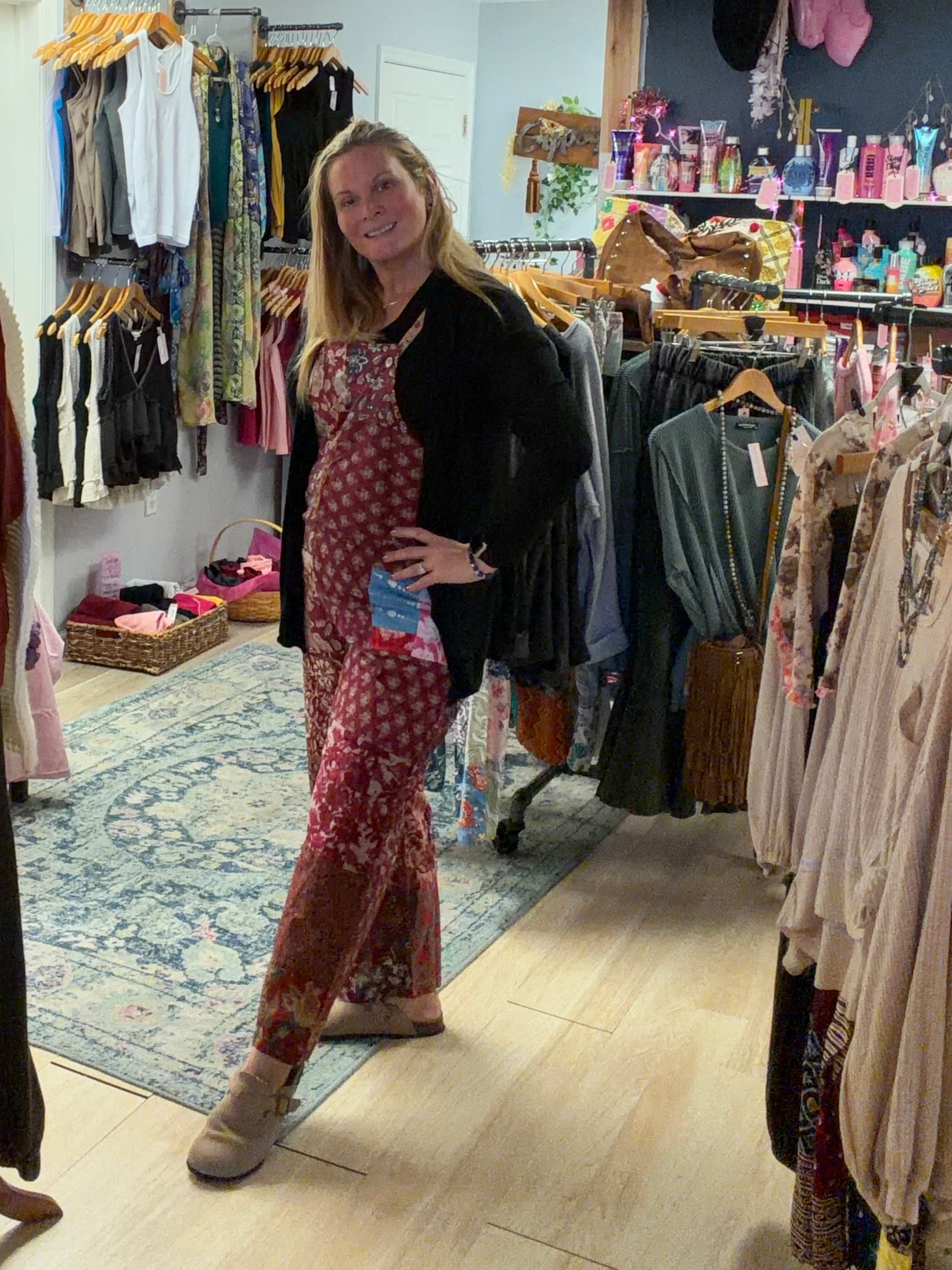 Woman wearing burgundy floral overalls with black long sleeve top standing in bohemian boutique in Berlin Maryland. Beach Gypsy boutique owner showcasing boho fashion styling for women who love free-spirited clothing and unique accessories.