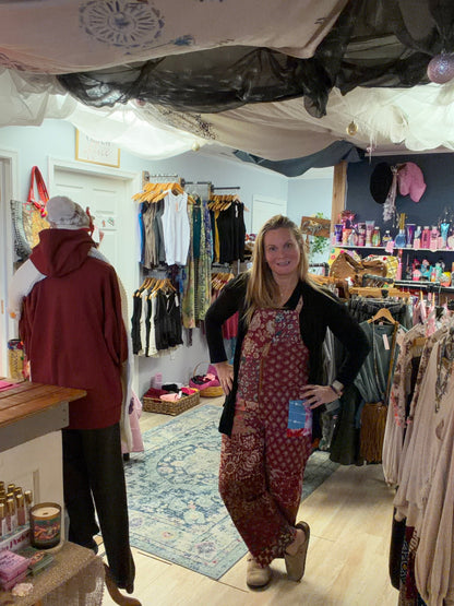 Woman wearing burgundy floral overalls with black long sleeve top standing in bohemian boutique in Berlin Maryland. Beach Gypsy boutique owner showcasing boho fashion styling for women who love free-spirited clothing and unique accessories.