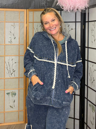 A woman wearing a matching dark blue acid-wash lounge set with white trim stands in front of decorative folding screens. She is smiling and posing, with a pink feather boa and floral drapery hanging in the background.