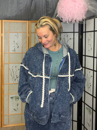 A woman wearing a blue denim-style cropped jacket with white trim over a green top, standing indoors in front of decorative folding screens and a draped backdrop with a pink feather pom-pom hanging overhead.