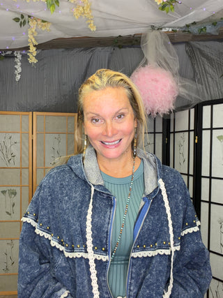 A smiling blonde woman wearing a blue denim hooded jacket with white lace trim over a sage green top, standing indoors in front of folding screens and a pink feather boa hanging from the ceiling.