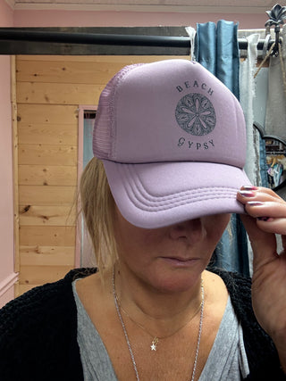Purple Beach Gypsy trucker hat on a woman standing in front of an indoor area with a wooden wall behind her 