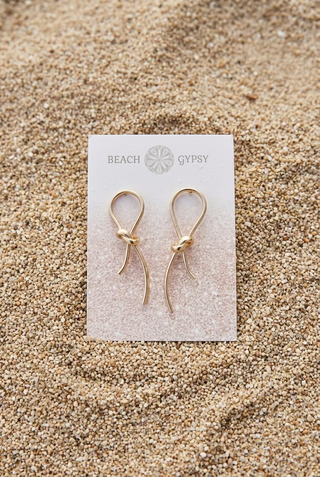 Gold Knot Post Earrings with a looped knot and dangling ends on a Beach Gypsy earring card, resting on beach sand