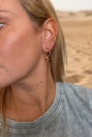 Woman wearing Gold Knot Post Earrings with a gray tee at the beach, a close-up of the gold knot bow earring