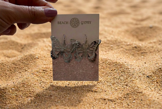 Silver Butterfly Earrings with open cutout wings on a Beach Gypsy earring card, held up against beach sand