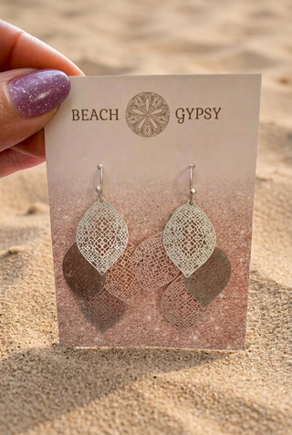 Silver-Tone Filigree Layered Leaf Dangle Earrings with lace-like openwork leaves on a Beach Gypsy earring card, held up against beach sand