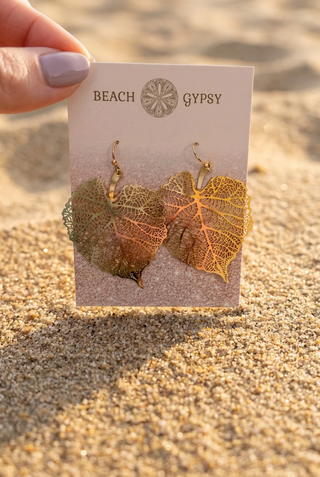 Gold Filigree Leaf Dangle Earrings with heart-shaped leaves and delicate vein detail on a Beach Gypsy earring card, held up against beach sand