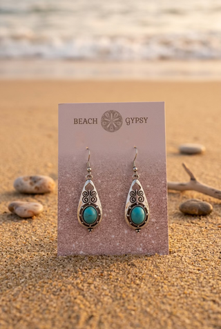 Turquoise-Color Scrollwork Teardrop Earrings with oval turquoise-color accents in silver-tone frames on a Beach Gypsy earring card, set on the sand at sunset by the ocean