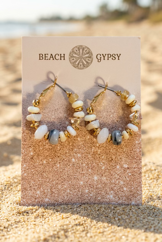 White and gray beaded hoop earrings with gold-tone accents on a Beach Gypsy earring card, set in the sand