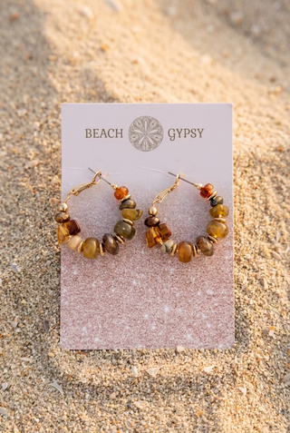 Amber and brown beaded hoop earrings with gold-tone accents on a Beach Gypsy earring card, set in the sand