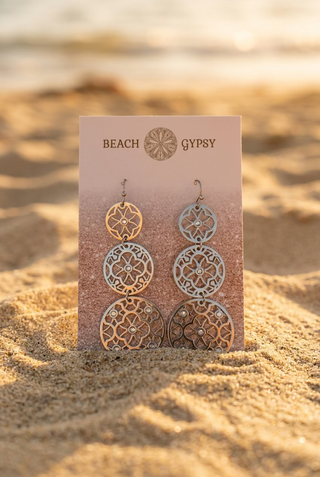 Silver Triple Floral Medallion Dangle Earrings with three graduated openwork flower medallions on a Beach Gypsy earring card, set in the sand at the beach