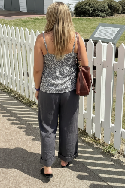 Womanwearing charcoal grey pants and a top  walking on a sidewalk with a white picket fence and grassy area in the background
