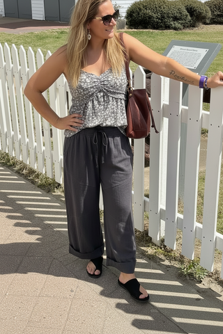 Woman standing outdoors near a white picket fence, wearing a patterned top and gray pants.