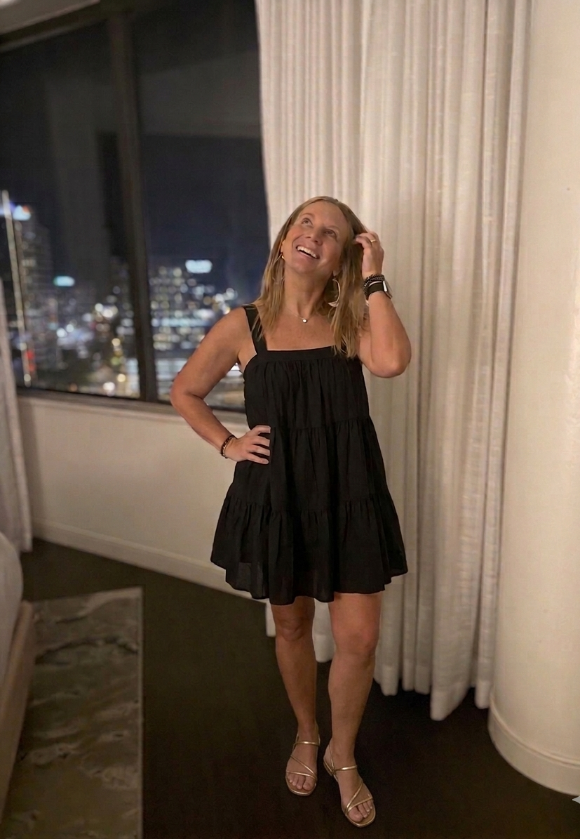 Woman in a black dress standing in a room with a cityscape view at night.