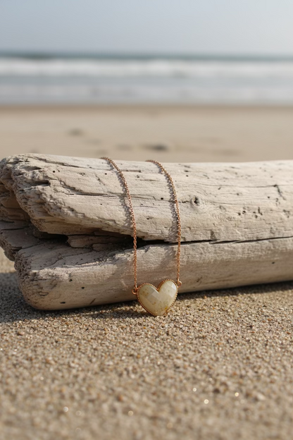 A gold heart-shaped pendant necklace with a textured, marbled surface and a small white inlay, hanging against a sandy background.