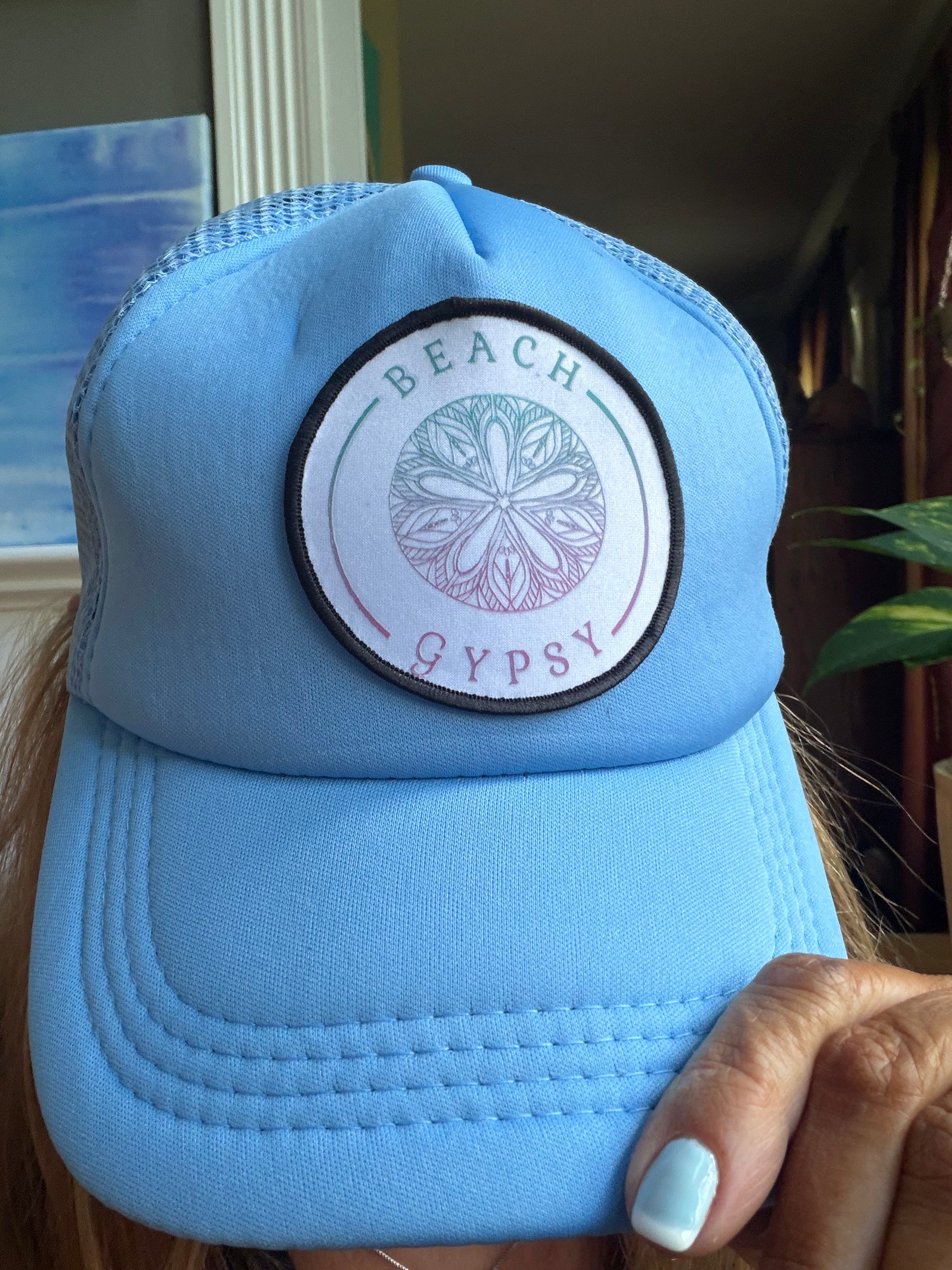 A light blue trucker hat with a circular 'Beach Gypsy' patch on the front, held by a hand in an indoor setting.